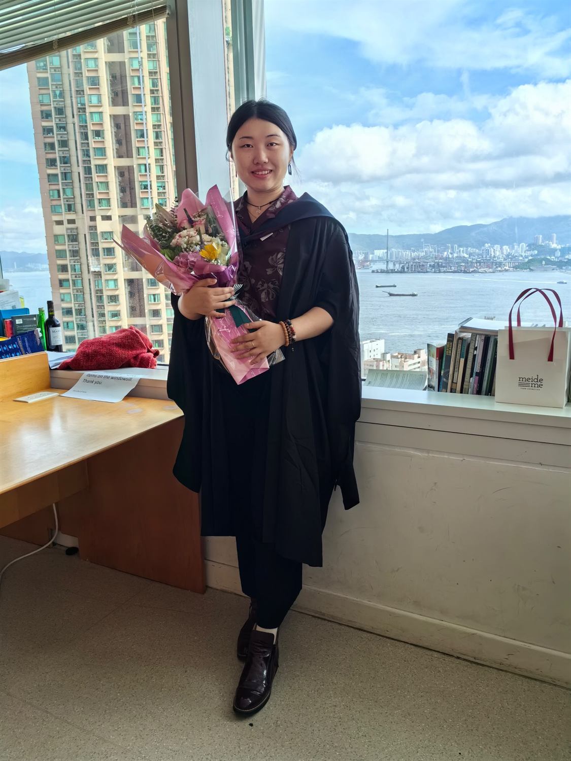 Zhixuan Li in graduation robes holding flowers, with Victoria Harbour behind, at the HKU Department of Philosophy